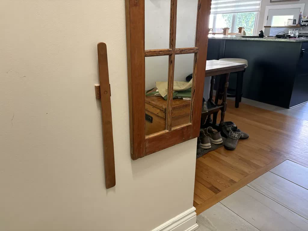 Chausse-pied en chêne clair sur un mur crème à côté d'un vieux miroir au cadre en bois brut, avec vue sur la cuisine