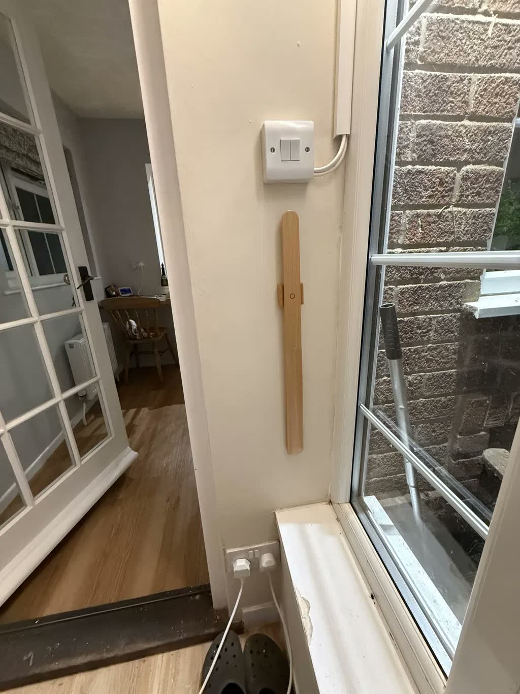 Chausse-pied en chêne clair sur un mur crème près d'une fenêtre et d'une porte, avec un interrupteur au-dessus et des sabots au sol